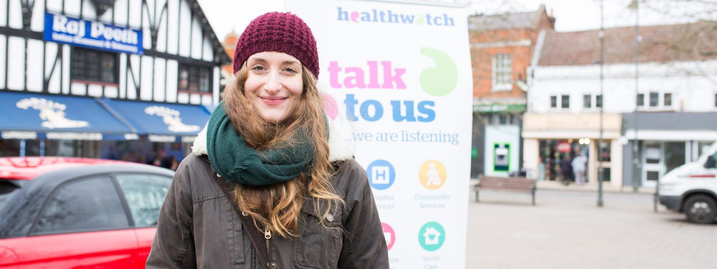 Young lady in a hat and scarf stood outside in front of a Healthwatch banner