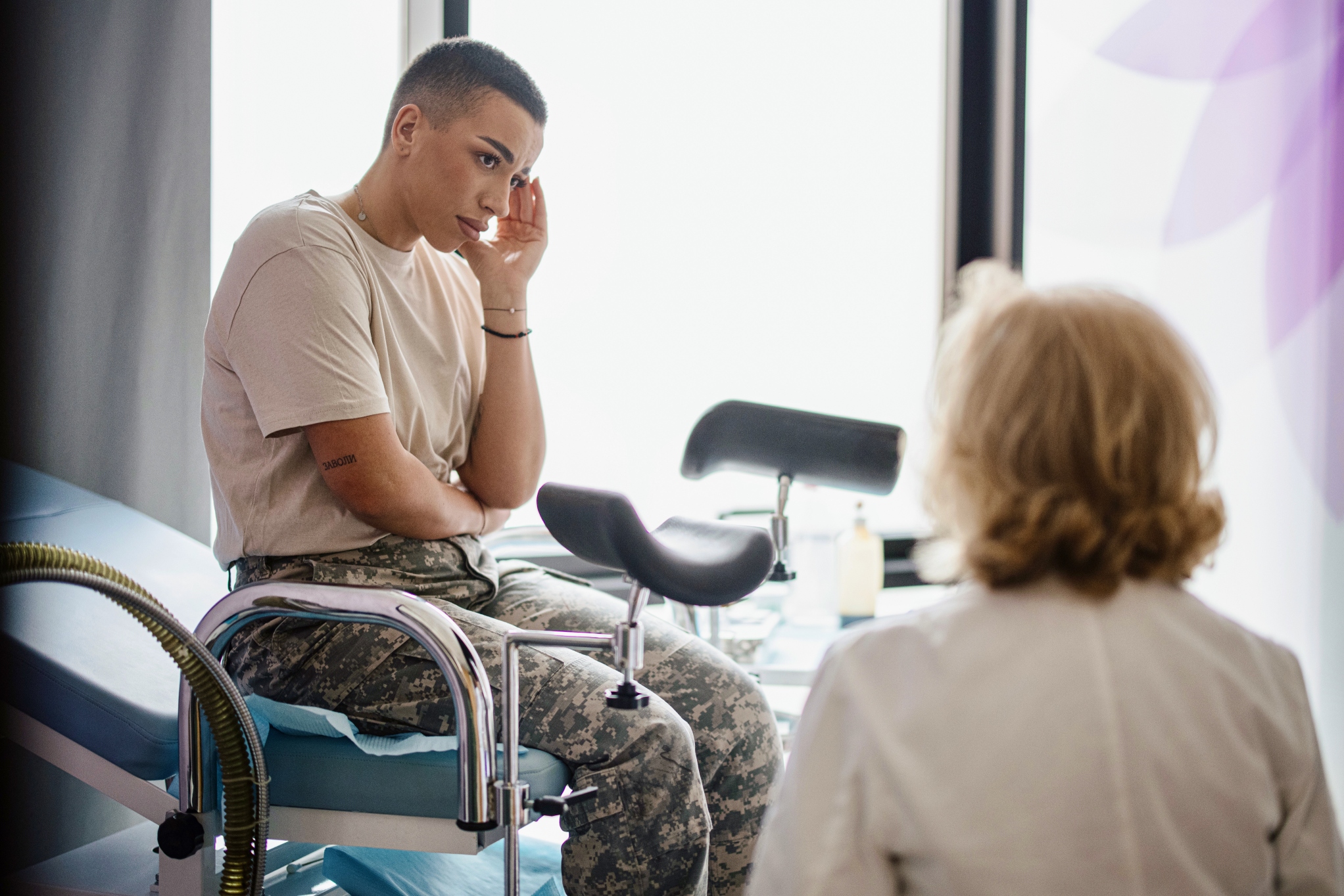 A woman wearing camouflage trousers sitting in a hospital bed, her hand on her cheek, talking to a doctor