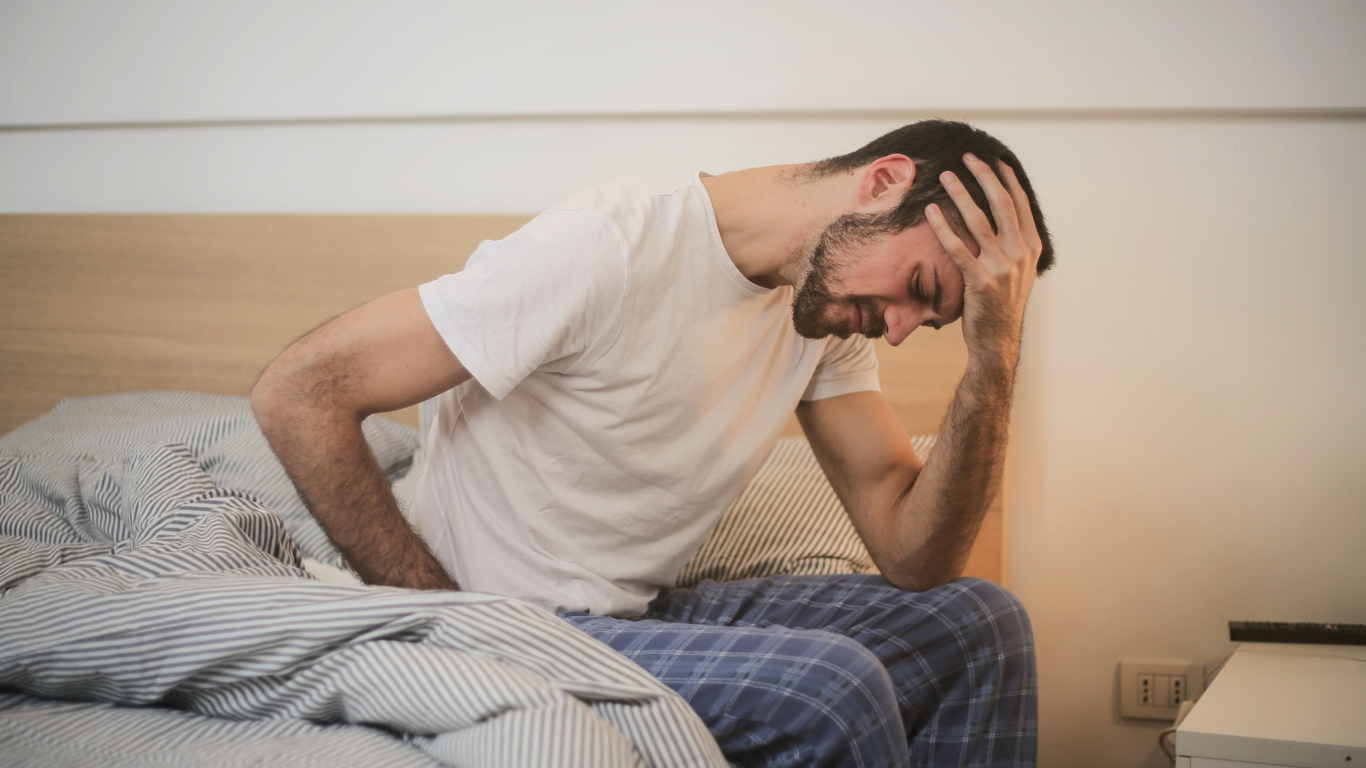 A man sits in a bed, holding his head