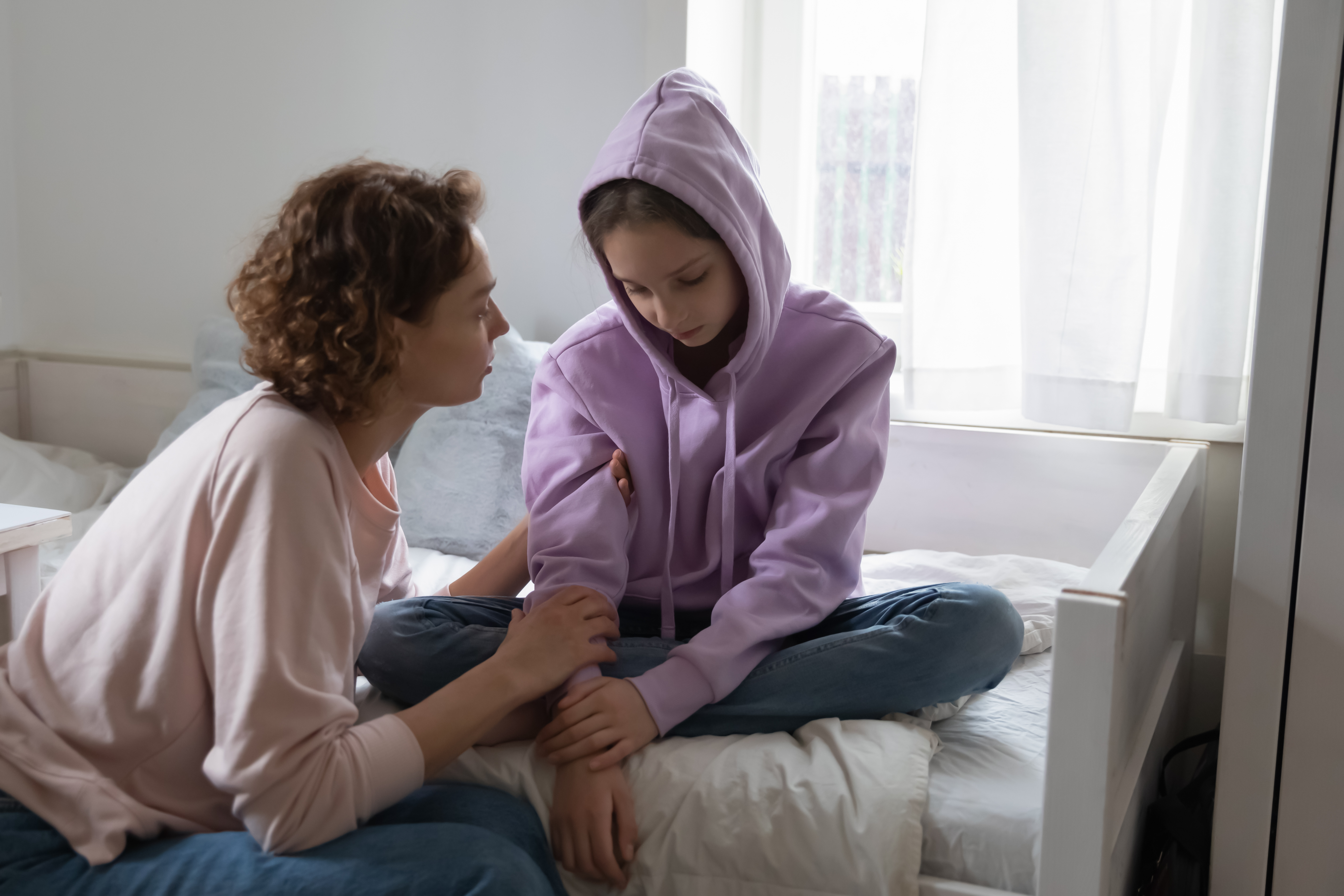 An upset child is sitting on a bed, while an adult leans over to console them