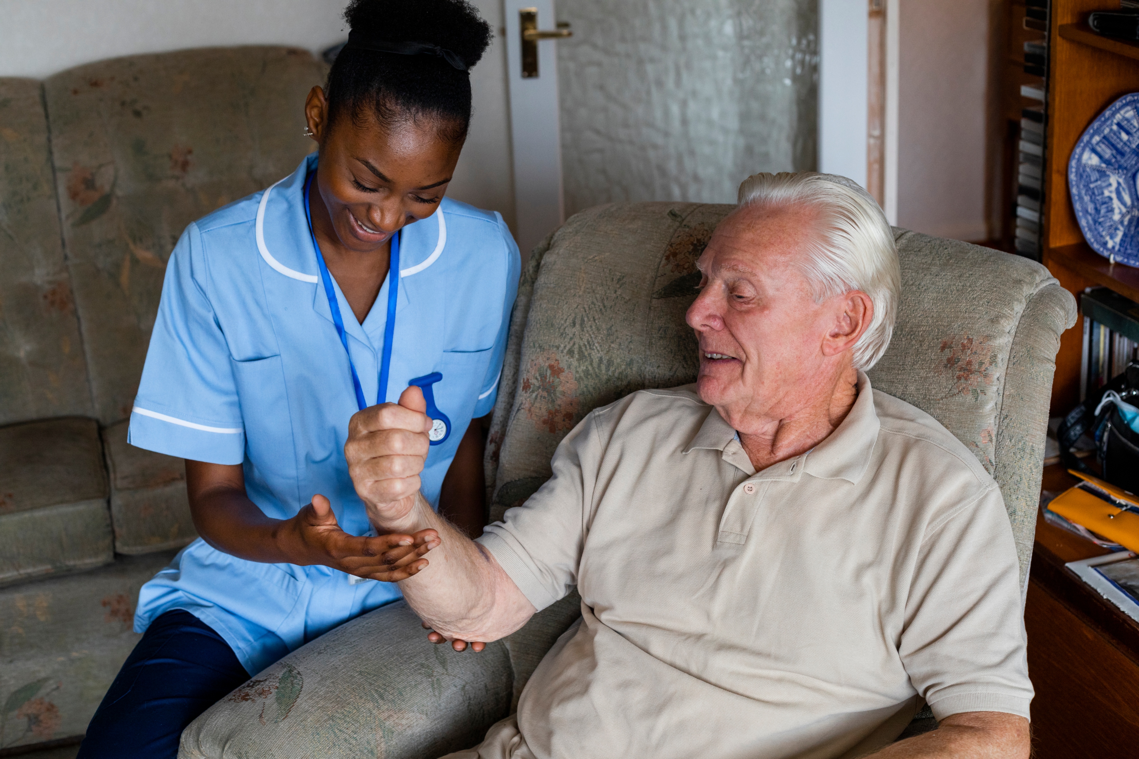 An elderly man sitting in a chair, while a carer holds his arm up slightly