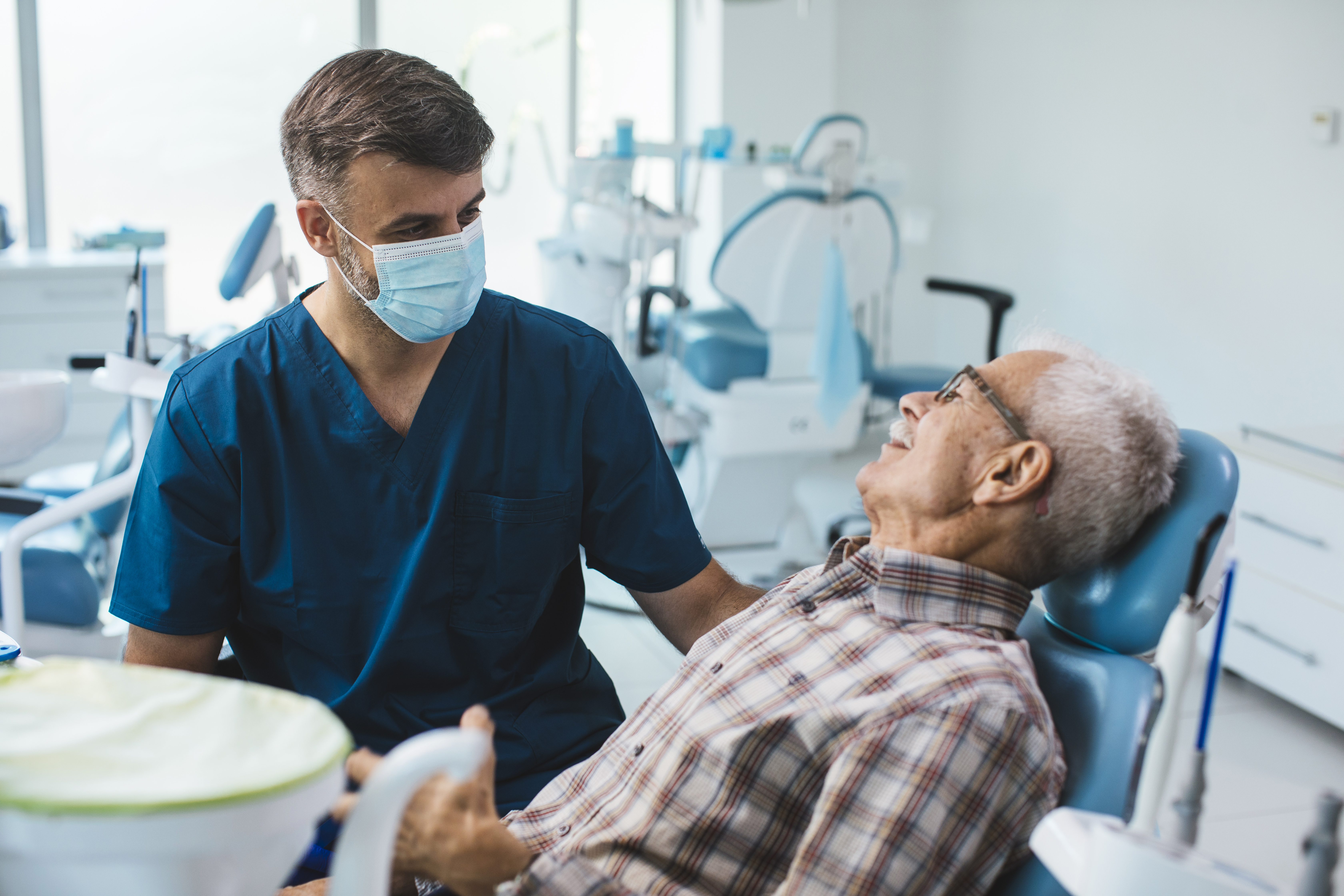 A dentist wearing a facemask speaks to an elderly man in a reclined chair