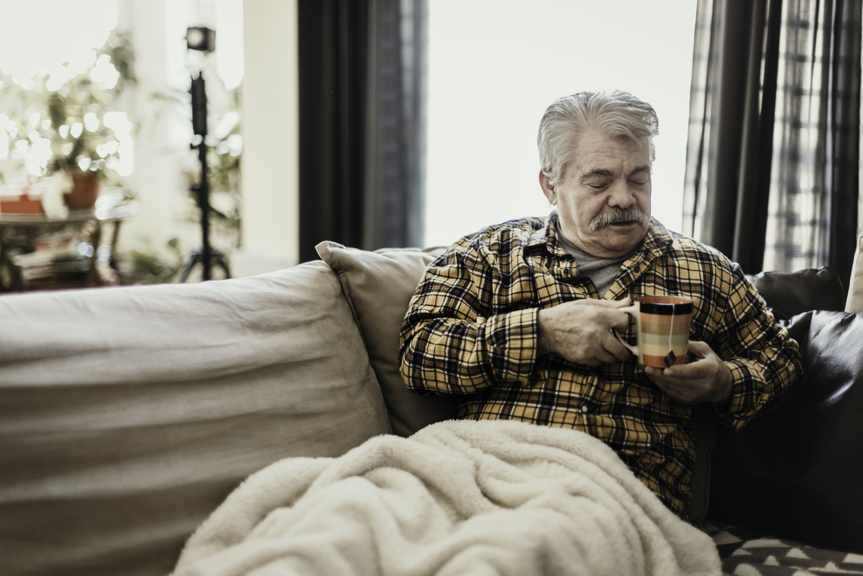 A man lies on a sofa with his legs under a blanket. He's holding a mug