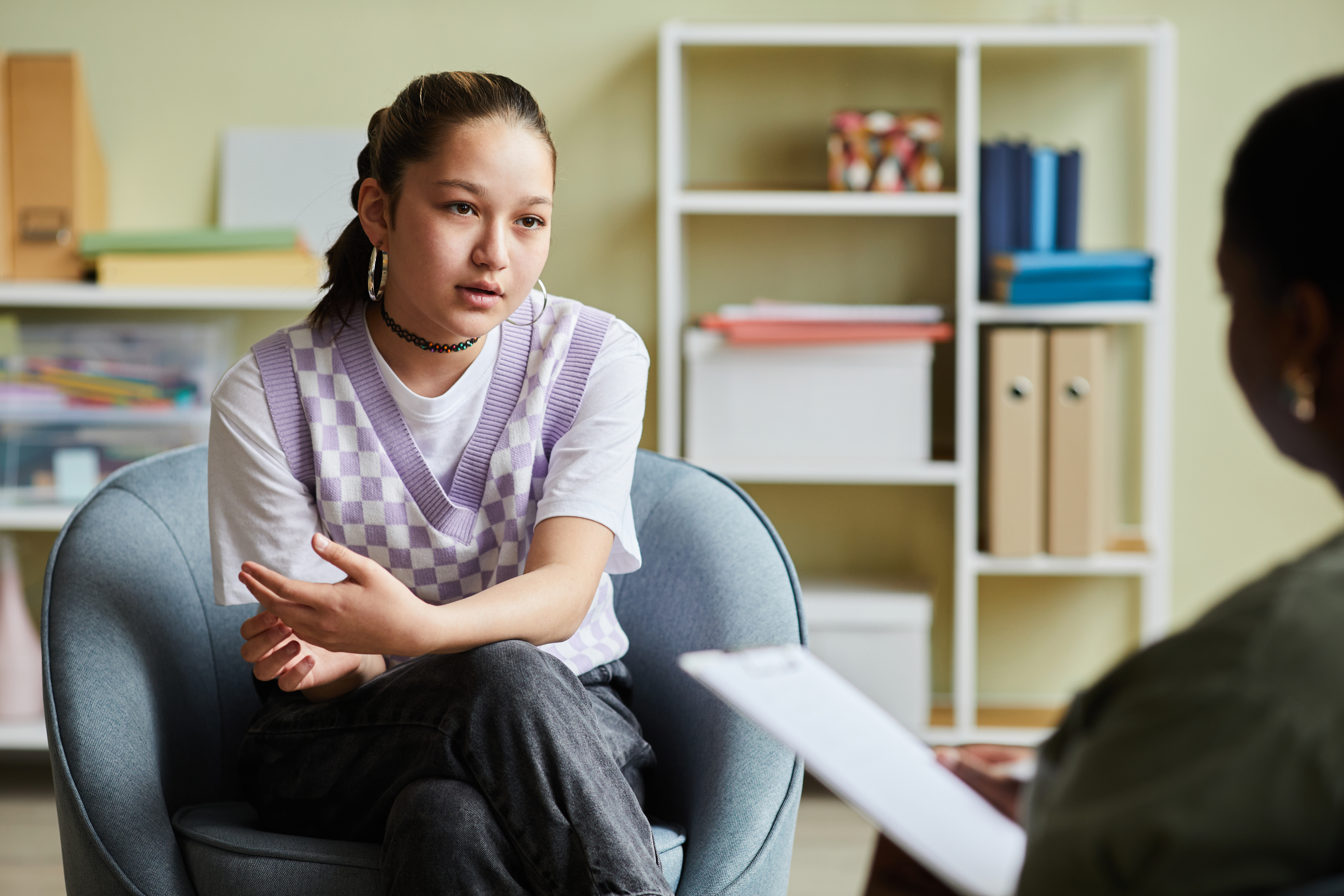 A young person sits in a chair leaning forwards, talking to an off-camera person holding a clipboard