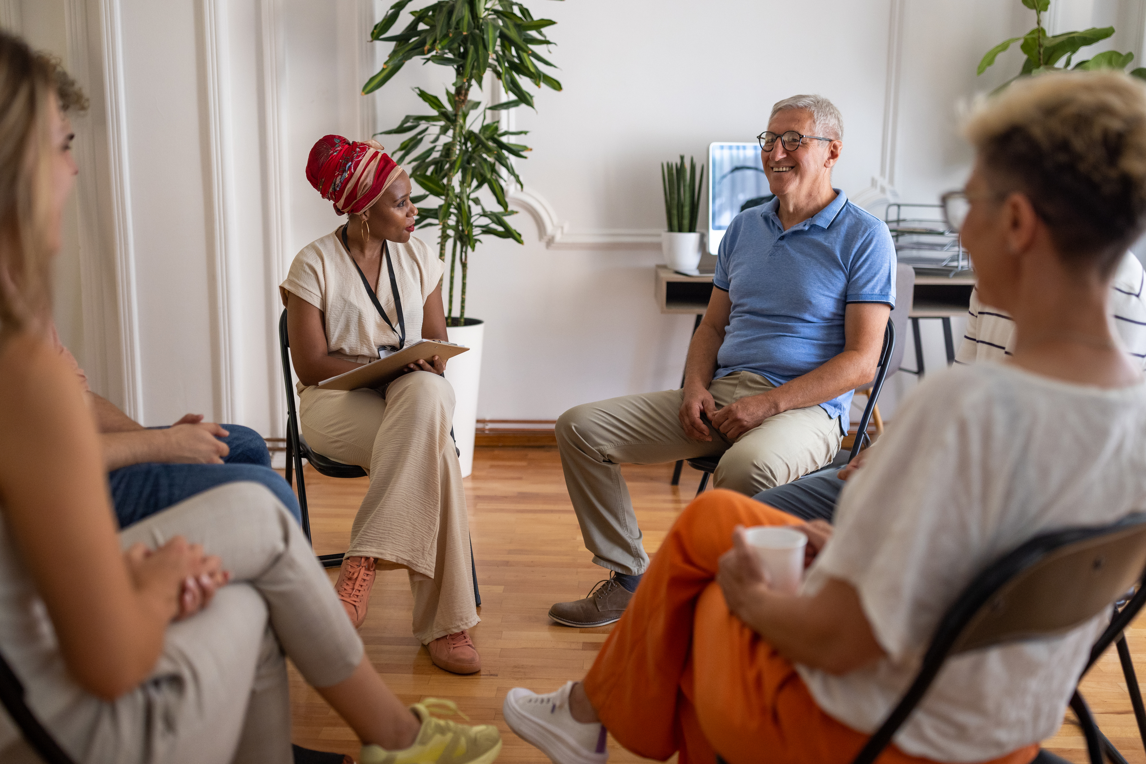 A group of four people sitting on chairs arranged in a circle. One woman is holding a clipboard