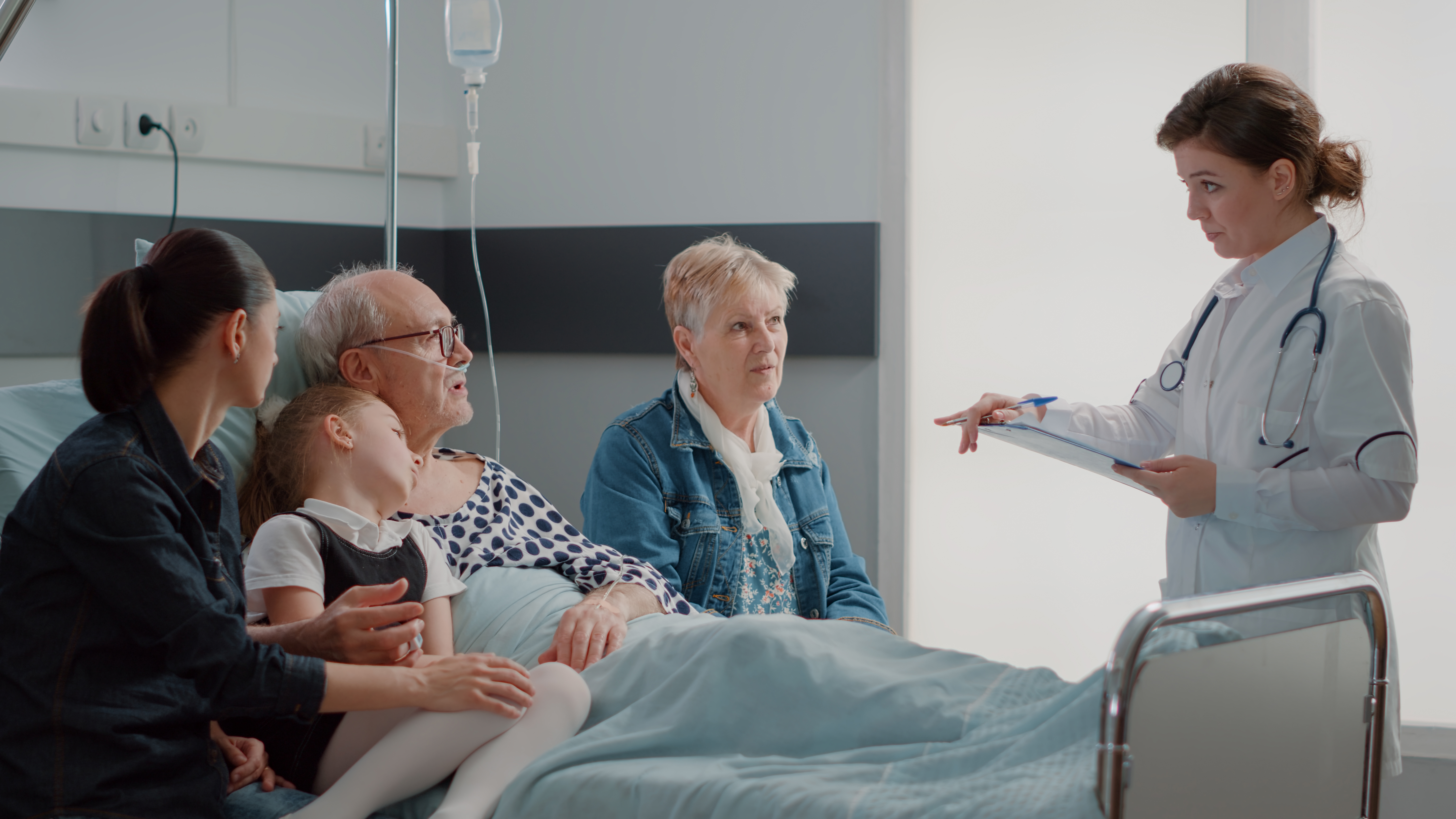 An elderly man in a hospital bed, with an adult and child sitting on one side and an elderly woman sitting on the other side, while a doctor stands at the foot of the bed with a clipboard
