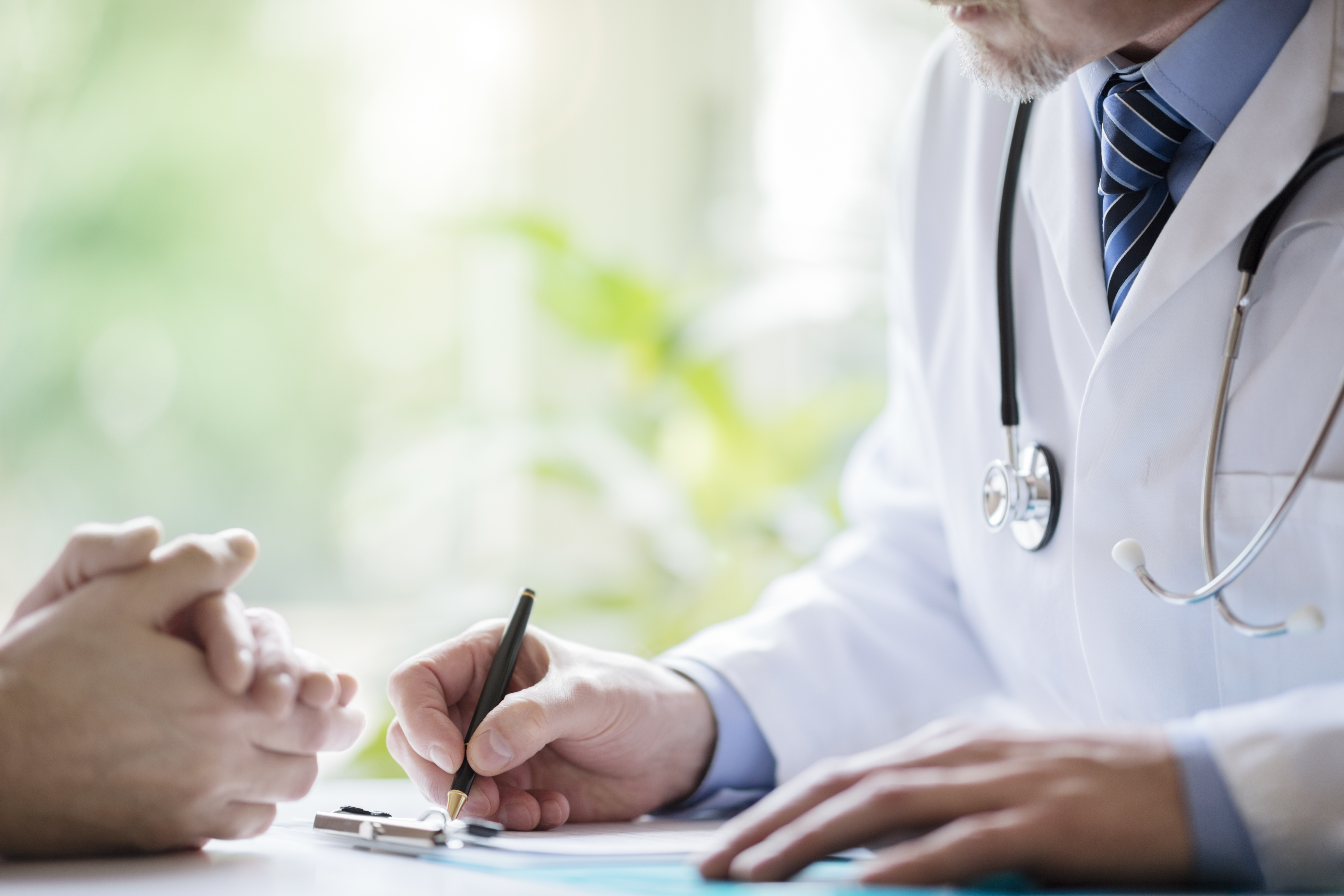 A close-up shot of a man wearing a labcoat and a stethoscope, writing something down on a piece of paper