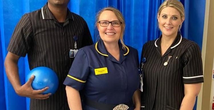 Three people in nursing and care uniforms, standing in a row and smiling for the camera. The man on the left is holding a ball