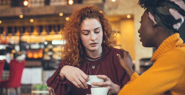 Two women having a conversation in a coffee shop. One has placed a hand on the other's shoulder reassuringly