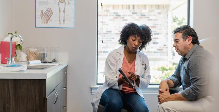 A man sits next to a woman in a GP's office. The woman is wearing a labcoat and is showing the man something on a tablet computer