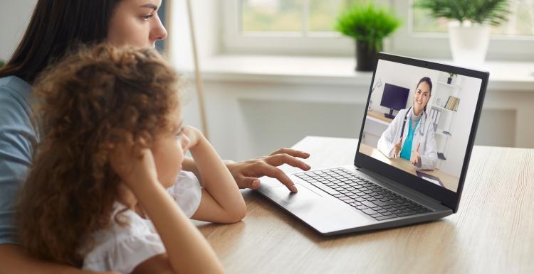 A woman and a child sit facing a laptop, which displays an image of a doctor