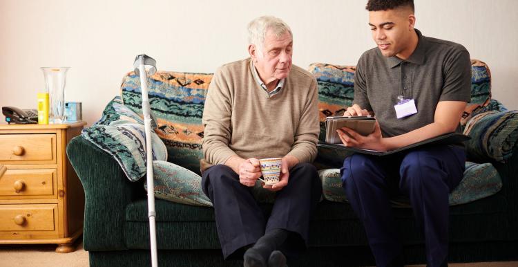 A carer sits on a sofa next to an elderly man, showing him something on a tablet
