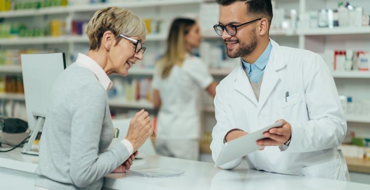 A woman speaks to a pharmacist over the counter, who is showing her a piece of paper