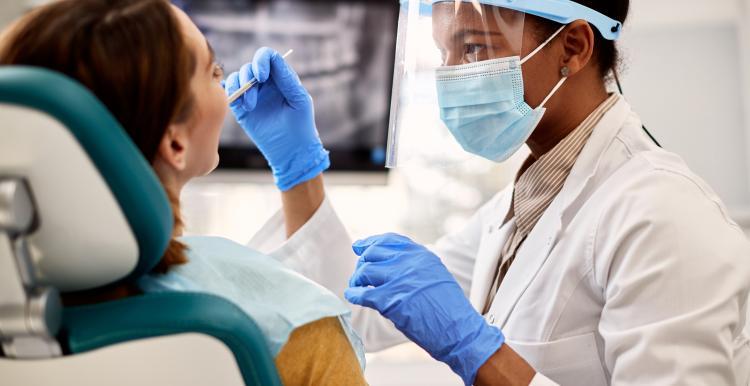 A dentist with a face mask, plastic face shield, and rubber cloves is reaching towards someone's mouth