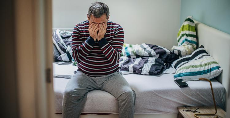 A man sits on an unmade bed, with his hands over his face