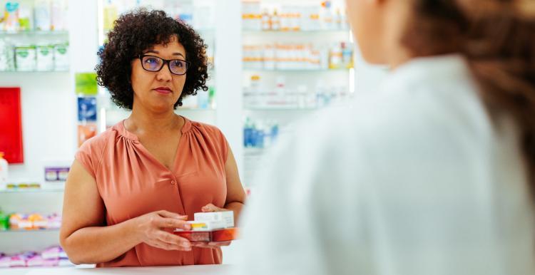 A pharmacist standing in front of shelves stocked with medicines, holding out several medication boxes