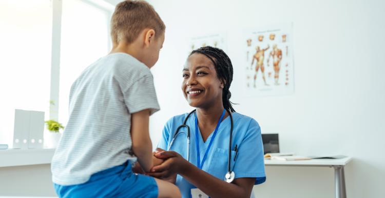 A child sits on a table, while a woman in a nurses' uniform sits in front of him smiling