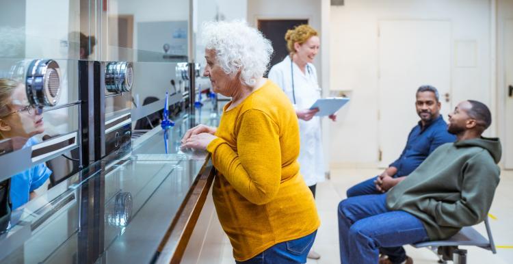 An elderly woman leans against a counter to talk to someone. In the background there are two people waiting, and a GP with a clipboard talking to them