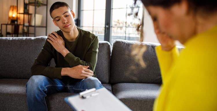 A young person sits on a sofa talking to someone, who is reading from a clipboard