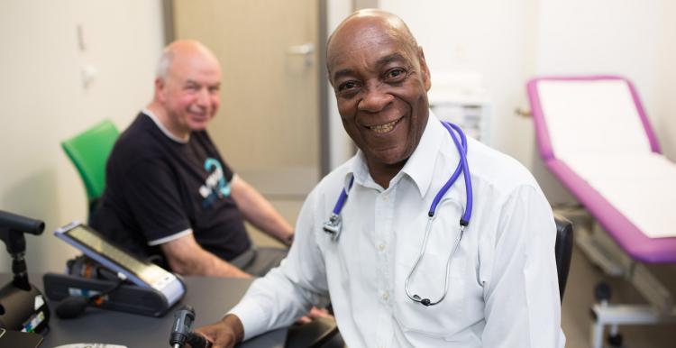 GP smiling at the camera with a patient in the background GP smiling at the camera with a patient in the background