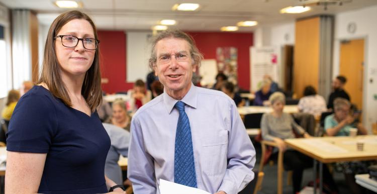 Man and a woman stood together holding some paperwork, in a busy room