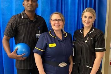 Three people in nursing and care uniforms, standing in a row and smiling for the camera. The man on the left is holding a ball