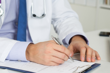 A man in a labcoat and wearing a stethoscope writes something on a clipboard