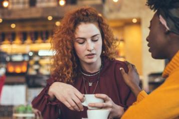 Two women having a conversation in a coffee shop. One has placed a hand on the other's shoulder reassuringly