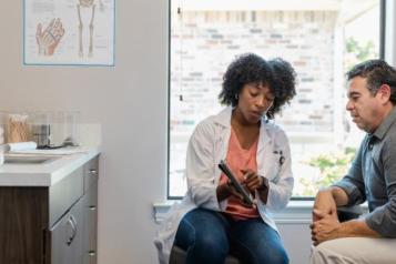 A man sits next to a woman in a GP's office. The woman is wearing a labcoat and is showing the man something on a tablet computer