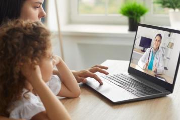 A woman and a child sit facing a laptop, which displays an image of a doctor