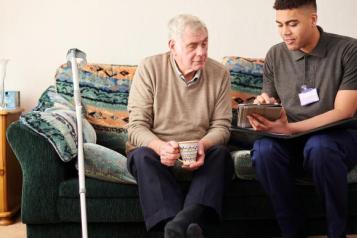 A carer sits on a sofa next to an elderly man, showing him something on a tablet