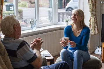 A woman sits on a footrest with a mug, talking to an elderly man in a chair