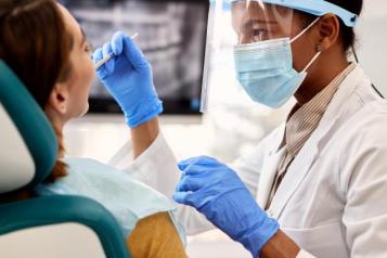 A dentist with a face mask, plastic face shield, and rubber cloves is reaching towards someone's mouth