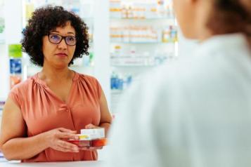 A pharmacist standing in front of shelves stocked with medicines, holding out several medication boxes