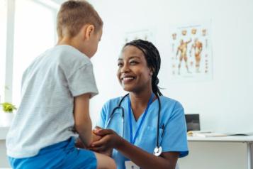 A child sits on a table, while a woman in a nurses' uniform sits in front of him smiling