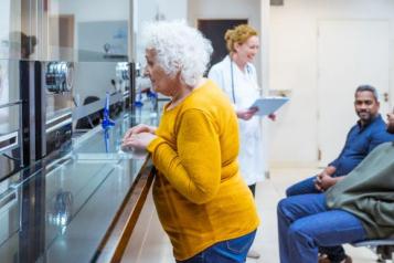 An elderly woman leans against a counter to talk to someone. In the background there are two people waiting, and a GP with a clipboard talking to them