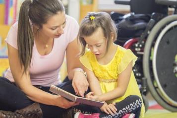 A woman sits on the ground with a child, who is reading from a book