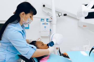 A dentist wearing a facemask leans over a patient in a reclined chair, performing a procedure on their mouth