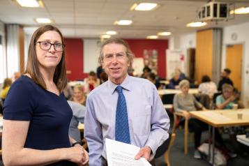 Man and a woman stood together holding some paperwork, in a busy room